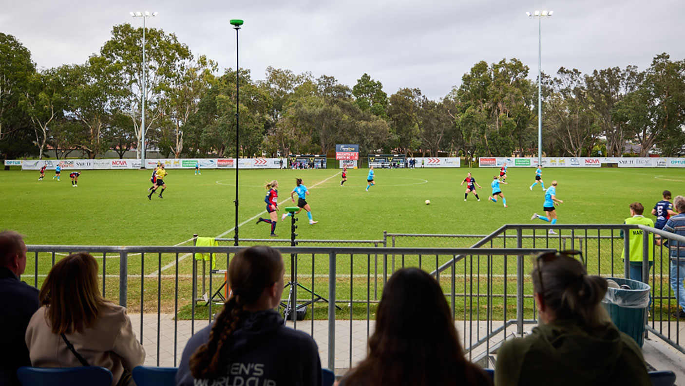 A soccer match on  grassy field, with an audience sitting in the stands in the foreground.